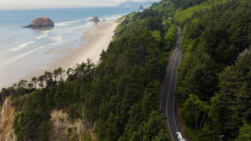 Drone aerial over Highway 101 on the Oregon Coast following cars driving along Arcadia Beach on a sunny summer afternoon with ocean waves and rocky shoreline.