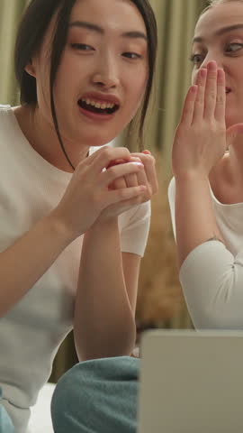 Two multiracial friends women talking gossip laughing with laptop on bed Caucasian woman whisper secret funny news to Asian Japanese Chinese Korean female girls friends spending time with computer