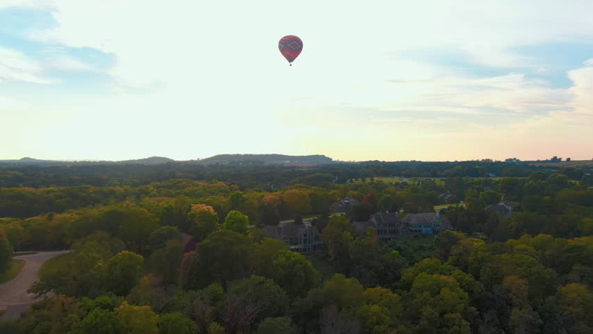 Aerial Pan-Up of a Lone Hot Air Balloon Floating Peacefully Over a Lush Forest with Affluent Neighborhood at Sunset