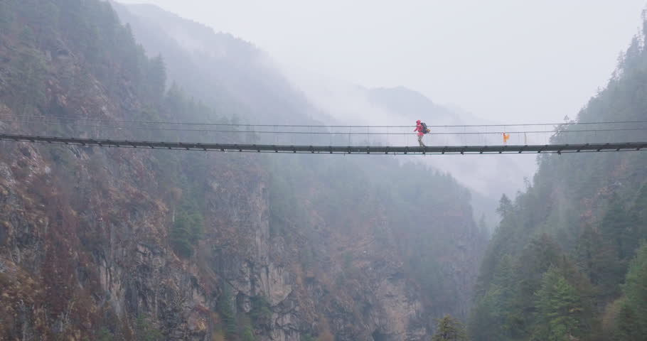 Drone view of Hillary Bridge near Monjo on Everest Base Camp trek, Nepal. A tall suspension bridge in rain shows tourists crossing, looking down at river and Himalayan landscape with awe and thrill