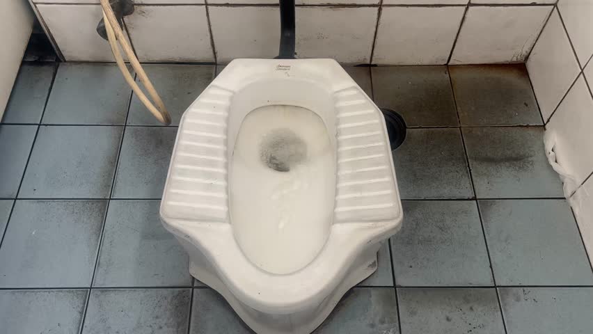 door in a public restroom. A short, vertical video of a dirty squat toilet flushing with water. The focus is on the mechanism of the flush and the unhygienic state of the restroom.