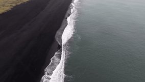 Aerial view of Reynisfjara's black sand beach, with waves rolling onto shore, Reynisdrangar sea stacks, green cliffs, and a nearby settlement. - Powered by Shutterstock - Get 15% off with code: PIKWIZARD15