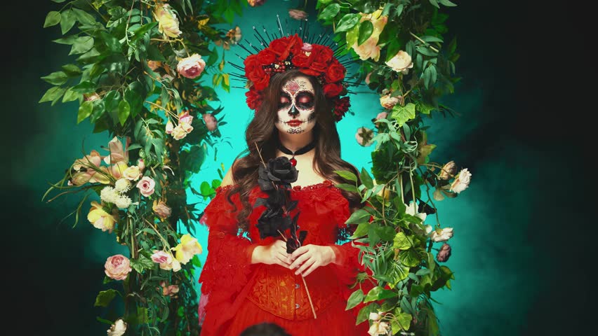 video real person gothic beauty woman sugar skull make-up art paint white face, hand holding black rose flower. red crown on head. Girl Calavera, day of the dead. dark night garden green trees arch