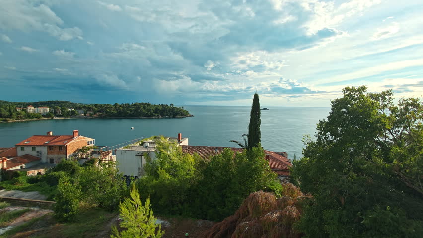 View Of Adriatic Sea From Rovinj Hill, Green Trees And Rooftops In Foreground, Calm Water And Dramatic Clouds In Distance, Peaceful Coastal Landscape