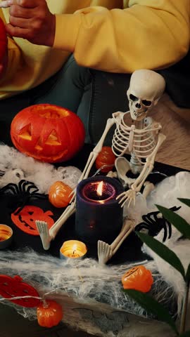 Girl in witch hat holding pumpkin in hand and painting spooky face in dark room with Halloween exposition on table. Halloween costume party. Vertical video