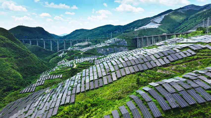 Aerial shot of a large solar farm with photovoltaic panels on green mountain, with a highway viaduct in the background.