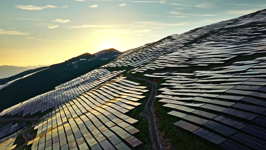 Aerial shot of a large solar power station on a mountainside, with panels reflecting the beautiful golden light of the rising sun.