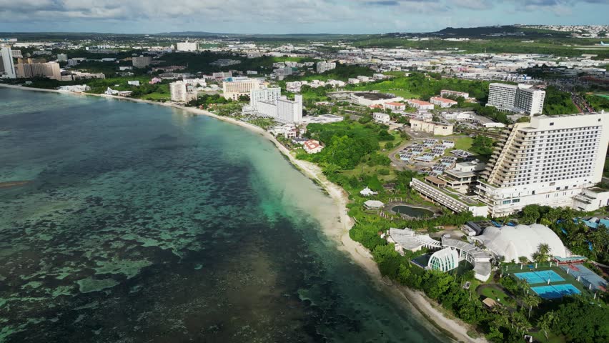 Stunning aerial flyover shot of vibrant shoreline and tropical beach resort and hotel buildings at Guam, USA.