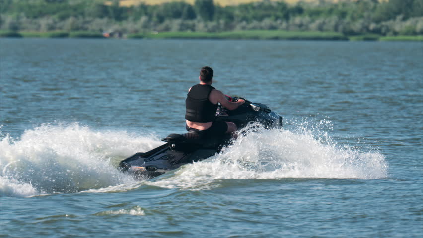 A man in a black life vest riding a black jet ski at high speed across a calm body of water