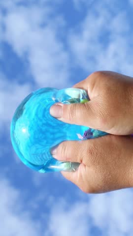 Hands firmly press the translucent blue slime of a jelly -like ball, filled with beads under daylight, the elastic surface is deformed by a noticeably playful movement. ASMR