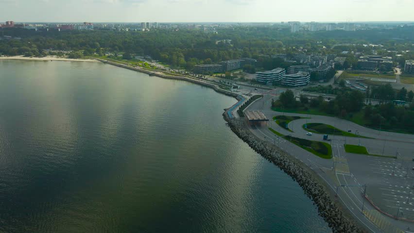 Aerial drone footage flying above deep dark water at a seaside shoreline in Pirita Estonia during a summer sunny day with a highway running next to the water where cars, vehicles and buses drive on.