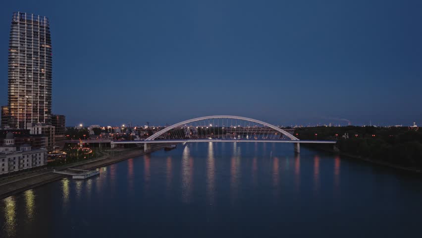 Aerial night view of the illuminated Apollo Bridge spanning the Danube River in Bratislava, Slovakia, with city lights reflecting on the calm water surface.