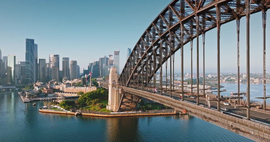 Australia, Sydney: Sydney Harbour Bridge in morning light. Cars driving traffic road connecting city skyline with surrounding urban landscape under bright blue sky. Aerial view drone flight footage