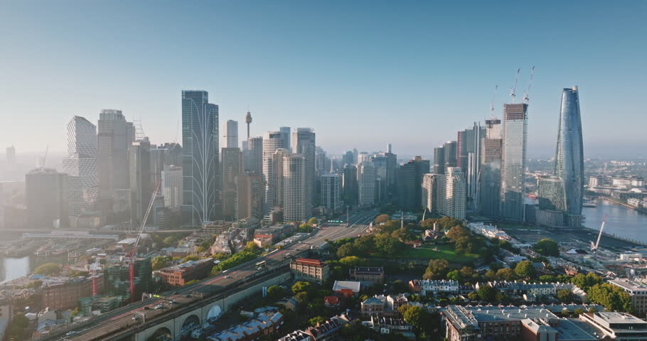 Australia, Sydney: Aerial view of Sydney Harbour Bridge and city skylines skyscrapers reflecting in water at misty sunrise, Barangaroo Reserve and wharves in foreground. Drone flight footage panorama