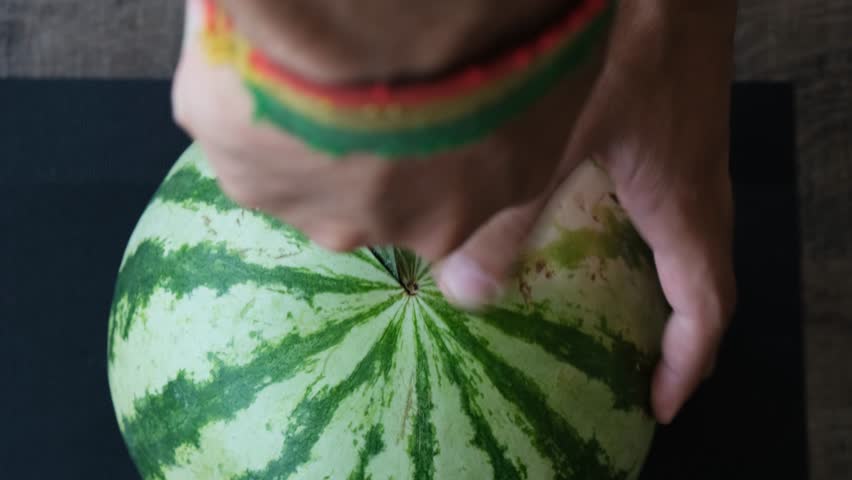 Top view of male hand using a large kitchen knife to cut a whole ripe striped watermelon into two halves. New harvest of fruits and berries. Watermelon is the largest berry by botanical definition.