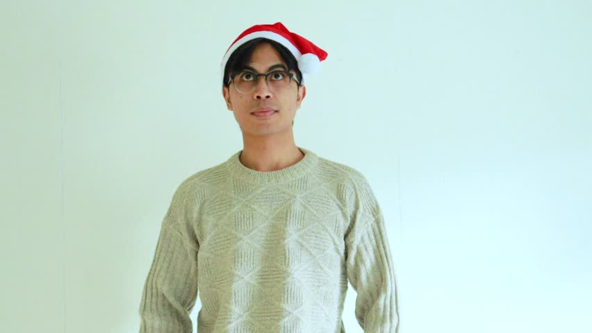 Excited young man wearing Christmas outfit and Santa hat pointing upward with joyful expression on studio background.