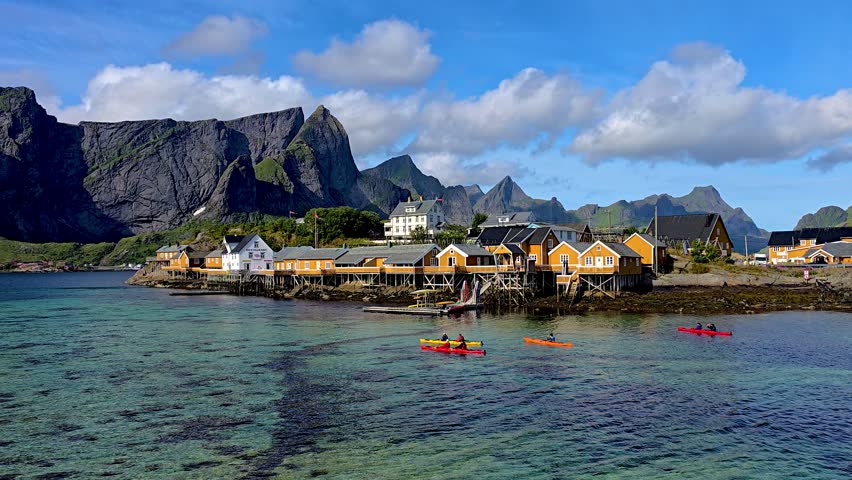 Colorful cabins line the shore as kayakers glide through clear waters in Lofoten, Norway. The majestic mountains rise dramatically, creating a breathtaking landscape perfect for outdoor adventures.