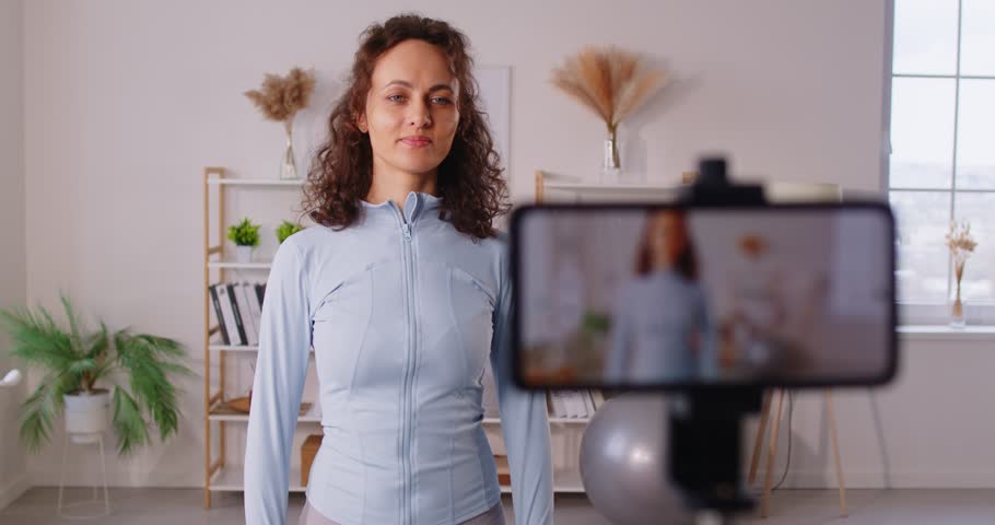 Sporty smiling young woman fitness instructor recording video and showing sports sneakers. Girl sport blogger making video for fitness blog on her smartphone about sportswear. 4k video.