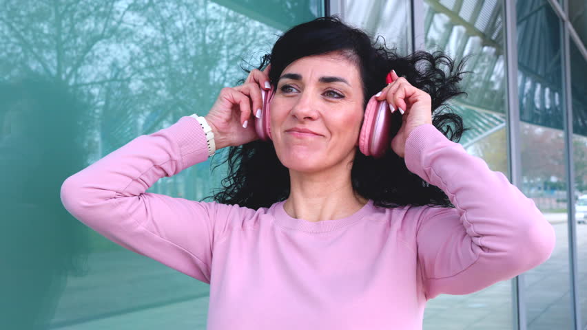 Smiling woman wearing pink headphones outdoors next to glass building, enjoying music and lifestyle, leisure activity, modern fashion, relaxation, audio, cheerful mood, casual urban environment