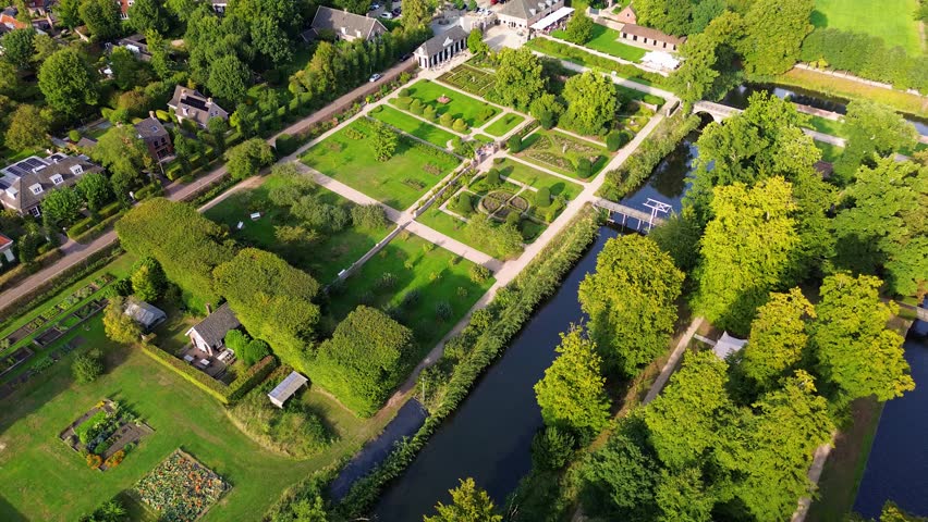 Aerial view of a formal garden with geometric layout, bordered by a canal and trees. A white manor stands at the center, surrounded by paths, hedges, and elegant landscaping.