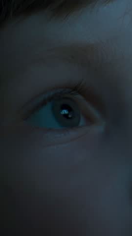 Close-up of a little boys eye, lit by the blue light of the cinema screen, with a reflection in his eye, as he watches intently and curiously at the movie on the TV, tablet, or plays computer games 
