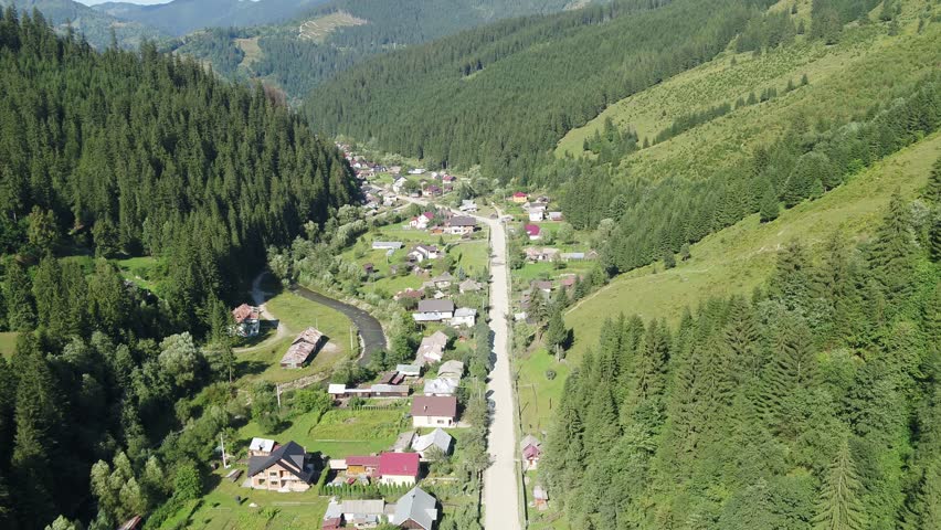 A high-angle aerial shot slowly orbits over a rural village in a Romanian mountain valley. Houses are scattered along a river and road, surrounded by lush green summer forests. Brosteni, Suceava.