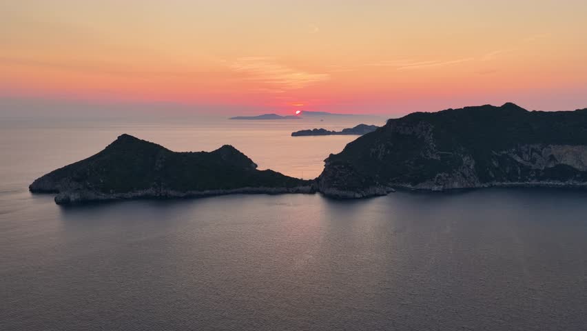 A stunning aerial view captures a breathtaking sunset over the calm, dark blue waters of the Ionian Sea. The sun dips below the horizon, casting a warm orange and pink glow across the sky.