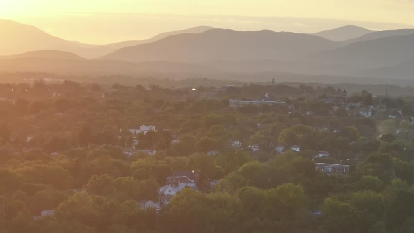 Cloudy sunrise in green American neighborhood with mountains in background. Suburb district with houses and green tress in quiet housing area. Aerial zoom wide shot.