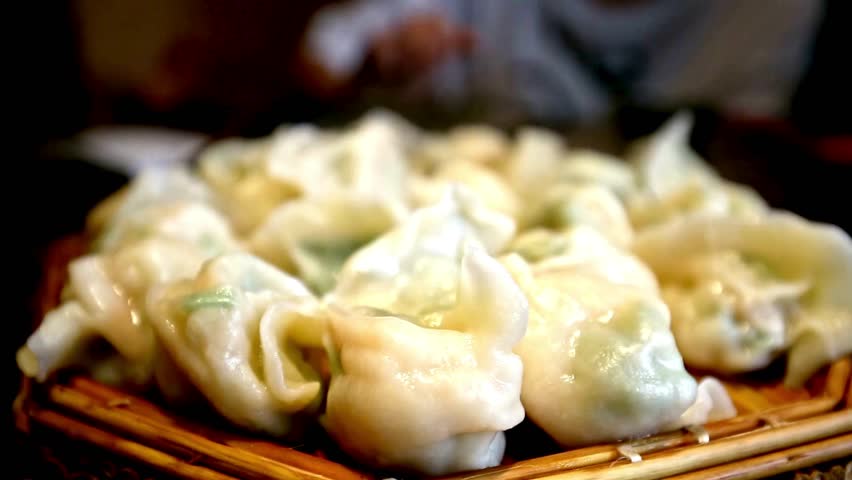 A close-up shot showcases a steaming platter of freshly made Asian dumplings, served traditionally in a woven bamboo basket, looking plump, juicy, and delicious.