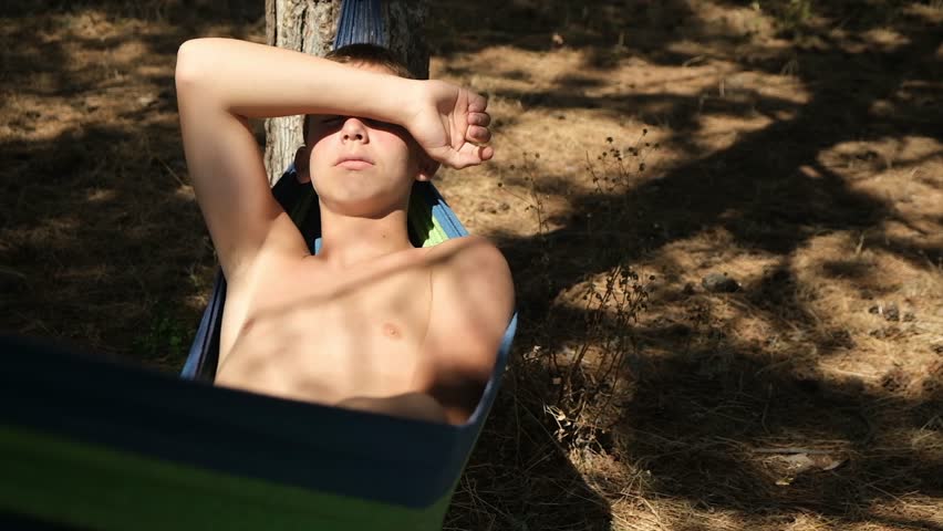 Young attractive shirtless man lies in a hanging hammock and covers his eyes from the sun with his forearm. The boy swings gently in the hammock outdoors among trees near the sea, resting in nature.