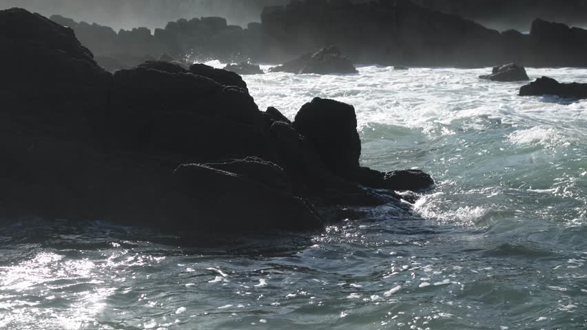 Waves collide with rocky shoreline, generating splashes and mist, illustrating the powerful forces of nature in a tranquil coastal setting