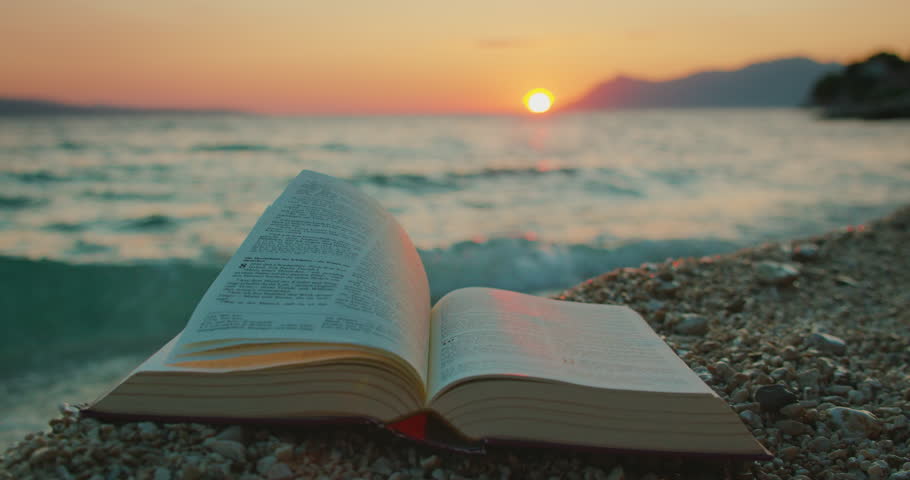 Open book Holy Bible rests on shore at sunset with sea and mountains in background