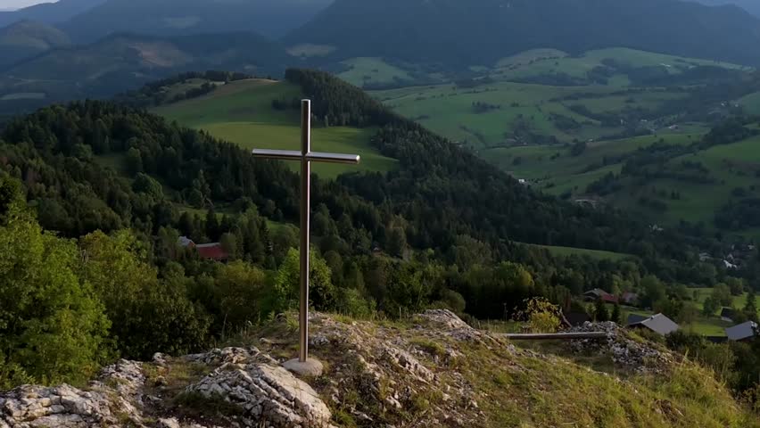 Cross on rocky hilltop overlooking green valley with forests and mountains in background