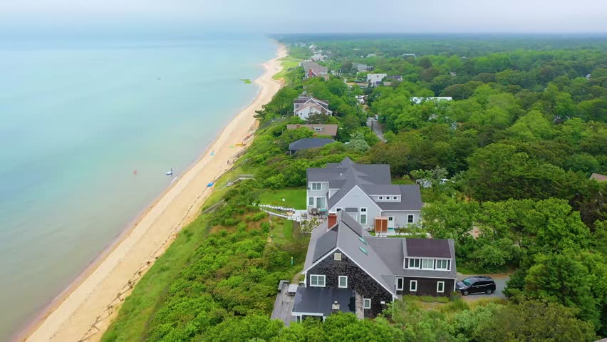 Scenic drone footage of Cape Cod coastal houses along a sandy beach, with cloudy skies overhead, calm Atlantic waters, and vibrant green forest neighborhoods behind the shoreline.