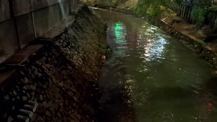 Urban river at night and dark concept. The river flows with reflections from lights and is illuminated by street lights, transportation and buildings. Urban activities and the river environment