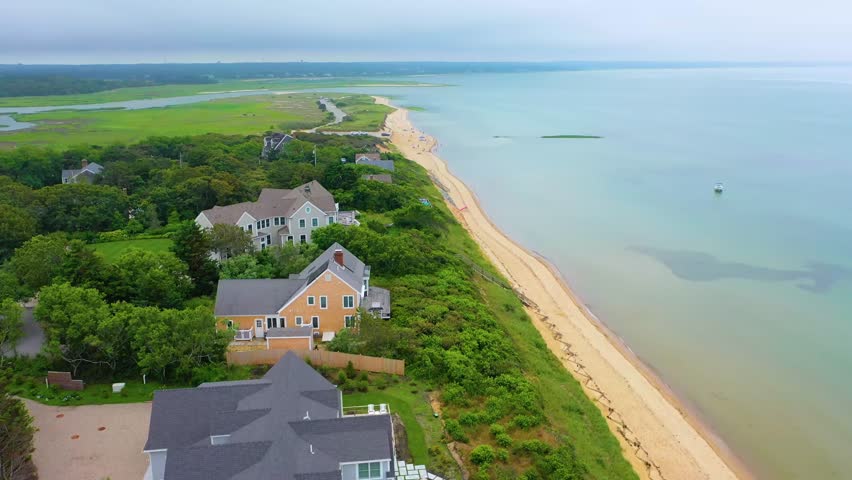 Drone panorama of Cape Cod shoreline featuring sandy beaches, coastal homes, and cloudy skies, with a sweeping view of neighborhoods and lush greenery meeting calm ocean waters.