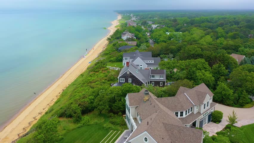 Scenic aerial panorama of Cape Cod beachside community, showcasing sandy shoreline, lush greenery, cloudy skies, and rows of oceanfront houses facing calm coastal waters.