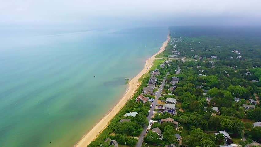 Drone footage of Cape Cod shoreline with oceanfront houses, sandy beach, cloudy skies, and lush green neighborhoods blending into the coastal landscape.