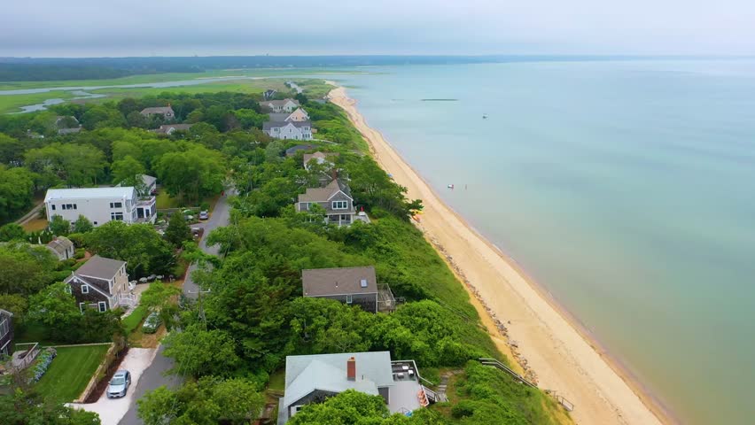 Aerial drone footage of Cape Cod coastline with beachfront houses, sandy shoreline, and cloudy skies stretching into the horizon above calm ocean waters and dense coastal neighborhoods.