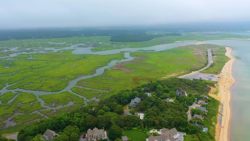 Cloudy day drone video of tidal marshes and sandy shoreline, with beach houses scattered among coastal trees, winding waterways feeding into the ocean, and calm waves breaking on the beach.