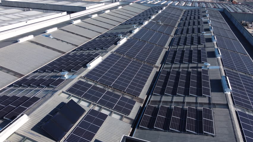 Drone footage flying over a large factory rooftop with long rows of solar panels. The perspective highlights renewable energy, industrial innovation and sustainable infrastructure.