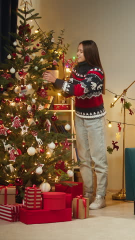 Woman decorating christmas tree. Young female student hanging balls and toys on evergreen tree in living room. Winter holidays concept, present box. Celebrating new year at home. 