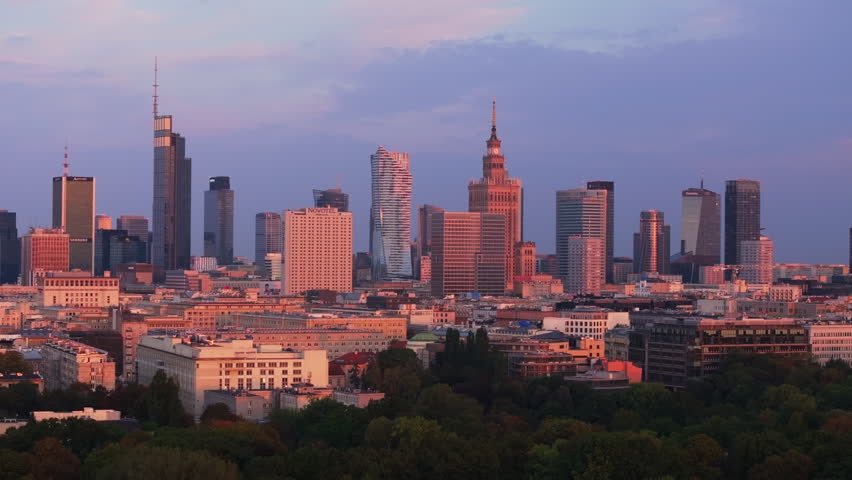 Modern skyscrapers of downtown Warsaw bathe in the warm light of the setting sun. Green trees cover a urban park just outside the financial district