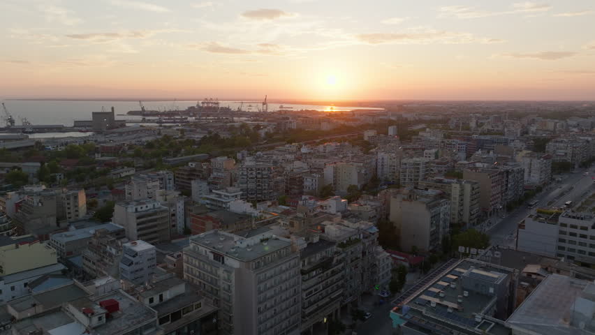 Aerial views capture Thessaloniki at sunset, with the sun setting over the Thermaic Gulf and casting a warm glow across the city, highlighting its vibrant skyline and architecture