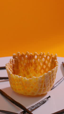 Empty waffle cone rotating surrounded by vanilla pods rotating against orange background. Vertical shot