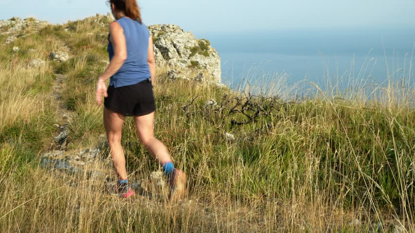 An adult woman dressed in sportswear walking along a path on a green cliff. The sea can be seen in the background. A summer day.