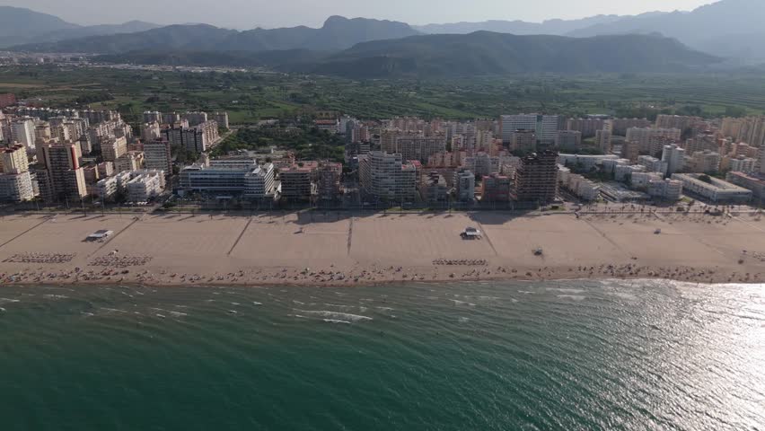 Aerial drone view of Gandia, Spain, showing sandy beach, city skyline, and mountains in the background on a sunny summer day