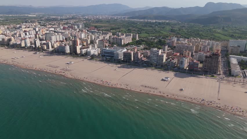 Aerial drone shot of Gandia, Spain, showing sandy beach, coastal buildings, and green mountains in the background on a sunny day