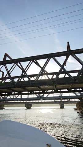 A first-person perspective from a motorboat, as it travels under a rusty steel railway bridge and a modern road bridge, with the bright sun reflecting off the water.