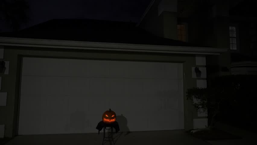 A carved jack-o-lantern glows on a stool in front of a closed garage door at night. Suburban driveway Halloween decoration with spooky light and shadows.